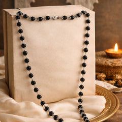 Black beaded necklace with silver clasps, draped over a beige fabric-covered box with a lit oil lamp in the background, creating a serene and elegant ambiance.