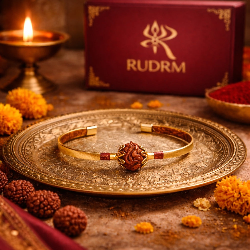 Rudraksha bracelet placed on an intricately designed golden plate, with marigold flowers and a lit oil lamp in the background, creating a warm and spiritual ambiance, alongside a 'RUDRM' box.