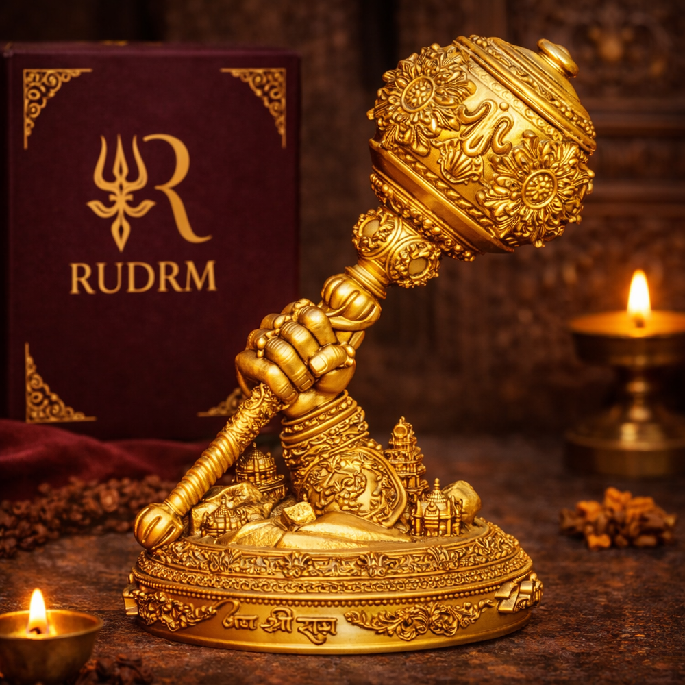 Golden intricately designed Hanuman Gada with detailed carvings, placed in front of a deep maroon box labeled 'RUDRM'. The scene is enhanced with two lit oil lamps and a decorative base with temple-like structures. The inscription 'Jai Shri Ram' is visible on the base of the gada.