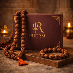 Rudraksha mala with a red tassel, placed beside a 'Rudrm' branded box, with a lit diya in the background.