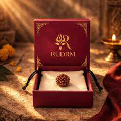 Rudraksha bead placed inside a maroon box with the 'RUDRM' logo, set against a background of lit oil lamps and marigold flowers, creating a serene spiritual atmosphere.