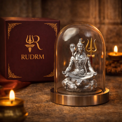 Silver statue of Lord Shiva under a glass dome, placed next to a 'Rudrm' branded box, with a lit diya and rudraksha beads in the background.
