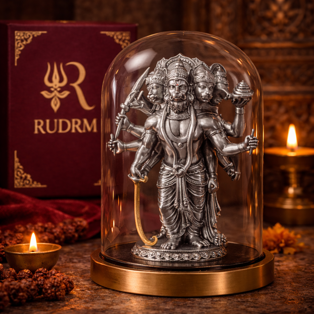 Silver statue of Lord Hanuman with multiple heads and arms under a glass dome, placed next to a 'Rudrm' branded box, with a lit diya and flower petals in the background.