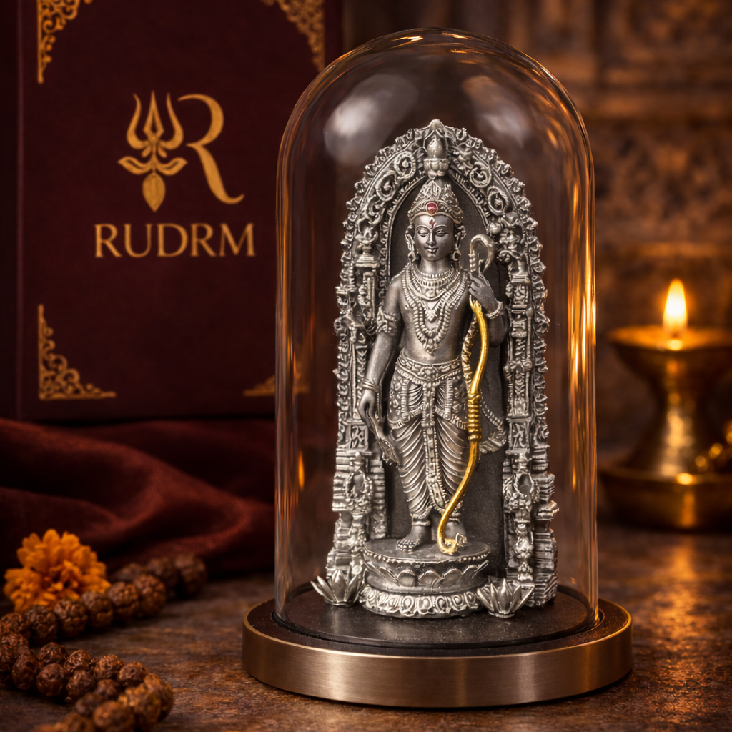 Silver statue of Lord Rama with a bow under a glass dome, placed next to a 'Rudrm' branded box, with a lit diya and rudraksha beads in the background.