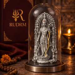 Silver statue of Lord Rama with a bow under a glass dome, placed next to a 'Rudrm' branded box, with a lit diya and rudraksha beads in the background.