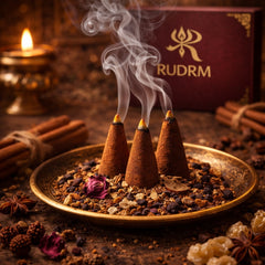 Three incense cones releasing aromatic smoke, placed on a brass plate with a mixture of dried flowers, spices, and herbs, alongside a lit candle and bundles of cinnamon sticks, with a Rudrm box in the background.