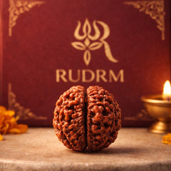 Close-up of a Rudraksha bead with intricate detailing, placed in front of a maroon 'RUDRM' box and a lit oil lamp, creating a spiritual and serene ambiance.