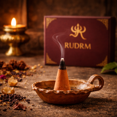Incense cone emitting smoke, placed in a traditional clay incense holder, with a 'Rudrm' branded box, dried herbs, and a lit diya in the background.
