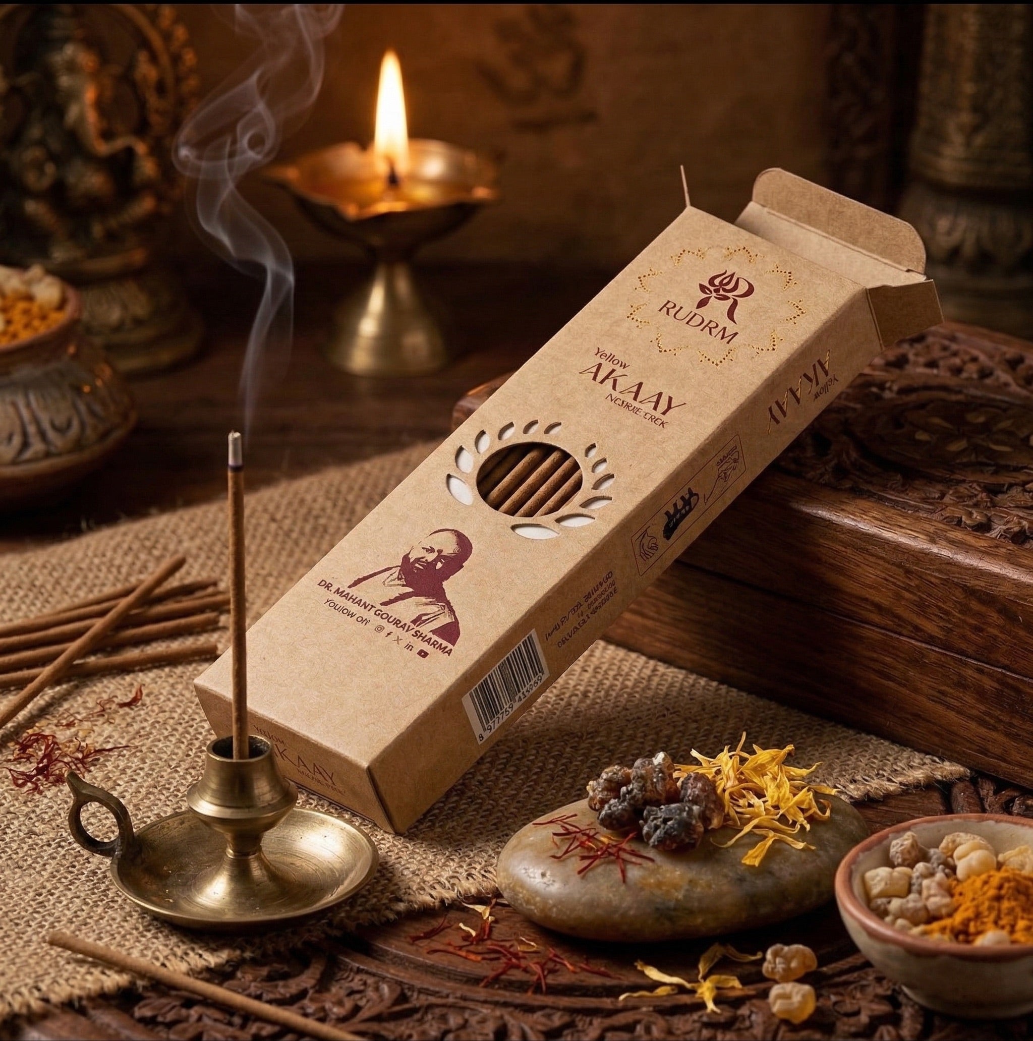 A box of Rudrm incense sticks with a portrait of Dr. Mahant Gourav Sharma on the packaging, placed on a wooden surface. The incense stick is burning in a brass holder, with a small plume of smoke rising. The background features a lit oil lamp, incense sticks scattered around, and a stone with aromatic spices like saffron and frankincense placed on it.
