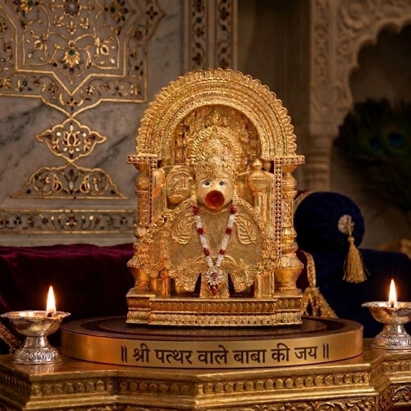 Golden statue of Lord Hanuman with a red nose, adorned with a rudraksha mala, placed on a pedestal with 'Shri Pathar Wale Baba Ki Jai' inscription, surrounded by lit diyas, set against an ornate background.