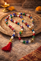 Colorful beaded mala with a red tassel, placed on a decorative metal plate, with a lit diya and marigold flowers in the background.