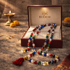 Colorful beaded mala with a red tassel, displayed in a 'Rudrm' branded box, with a lit diya and marigold flowers in the background.