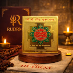 24K gold-plated Shri Narasimha Yantra with engraved mantras, placed on a wooden base, next to a 'Rudrm' branded box, with a lit diya and rudraksha beads in the background.