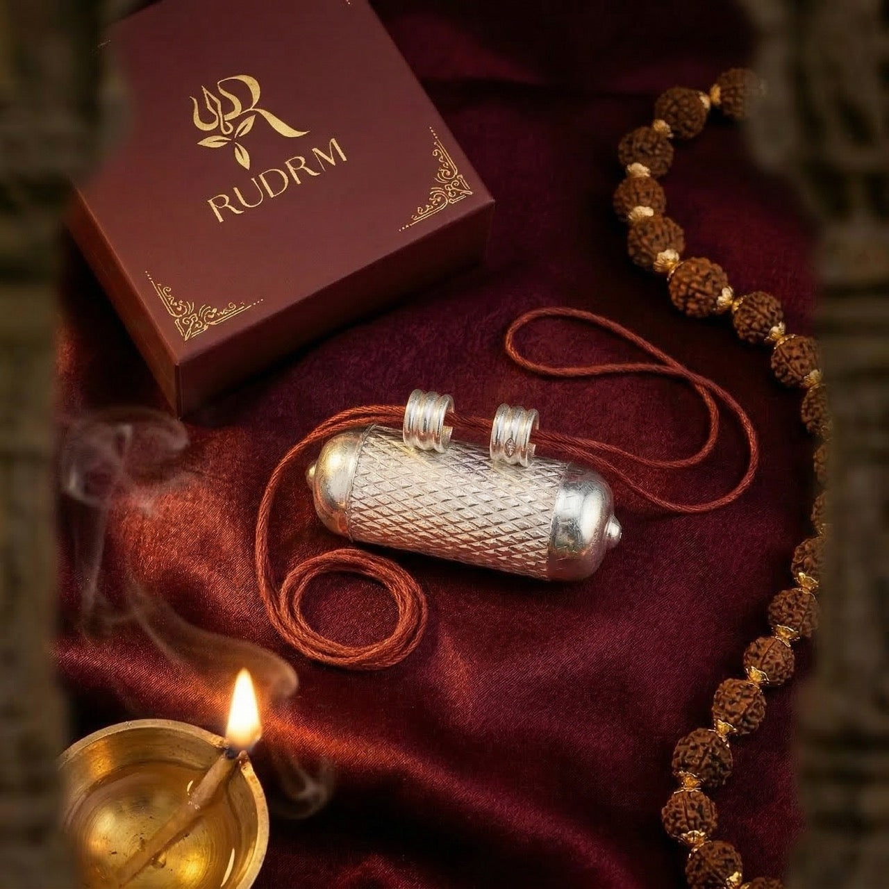 Silver Hanuman Gada pendant in a decorative box, with a rudraksha mala, incense smoke, and a lit diya on a maroon velvet background.