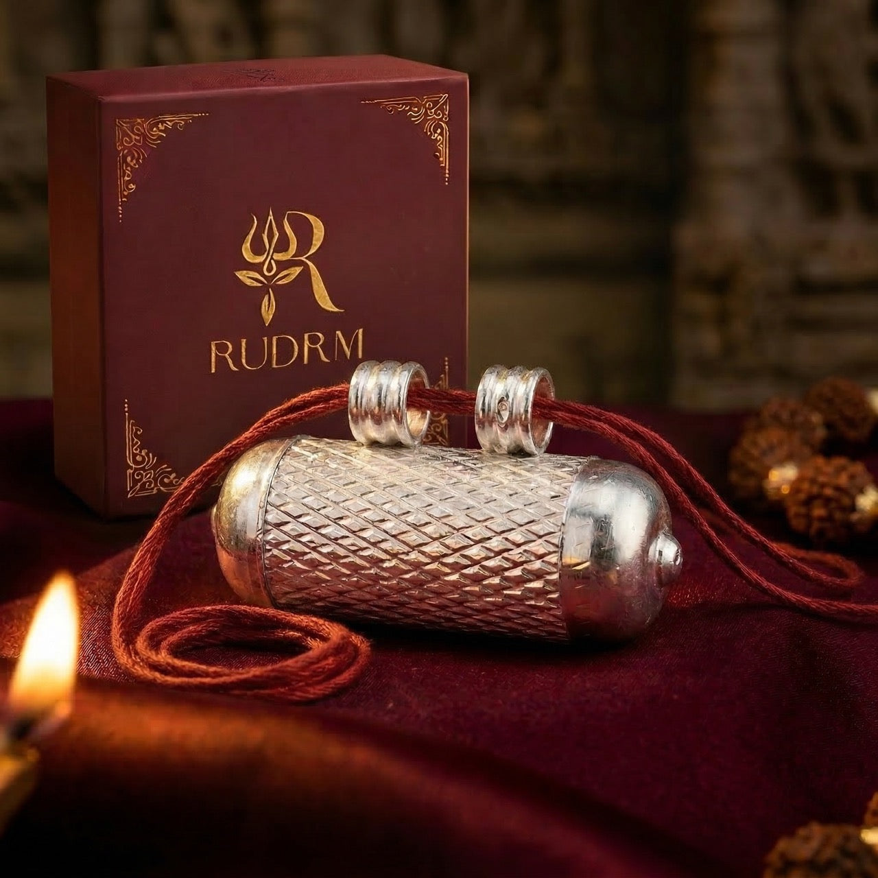 Silver Hanuman Gada pendant with red string, placed next to a 'Rudrm' branded box, set on a maroon velvet background with a lit diya and rudraksha beads.